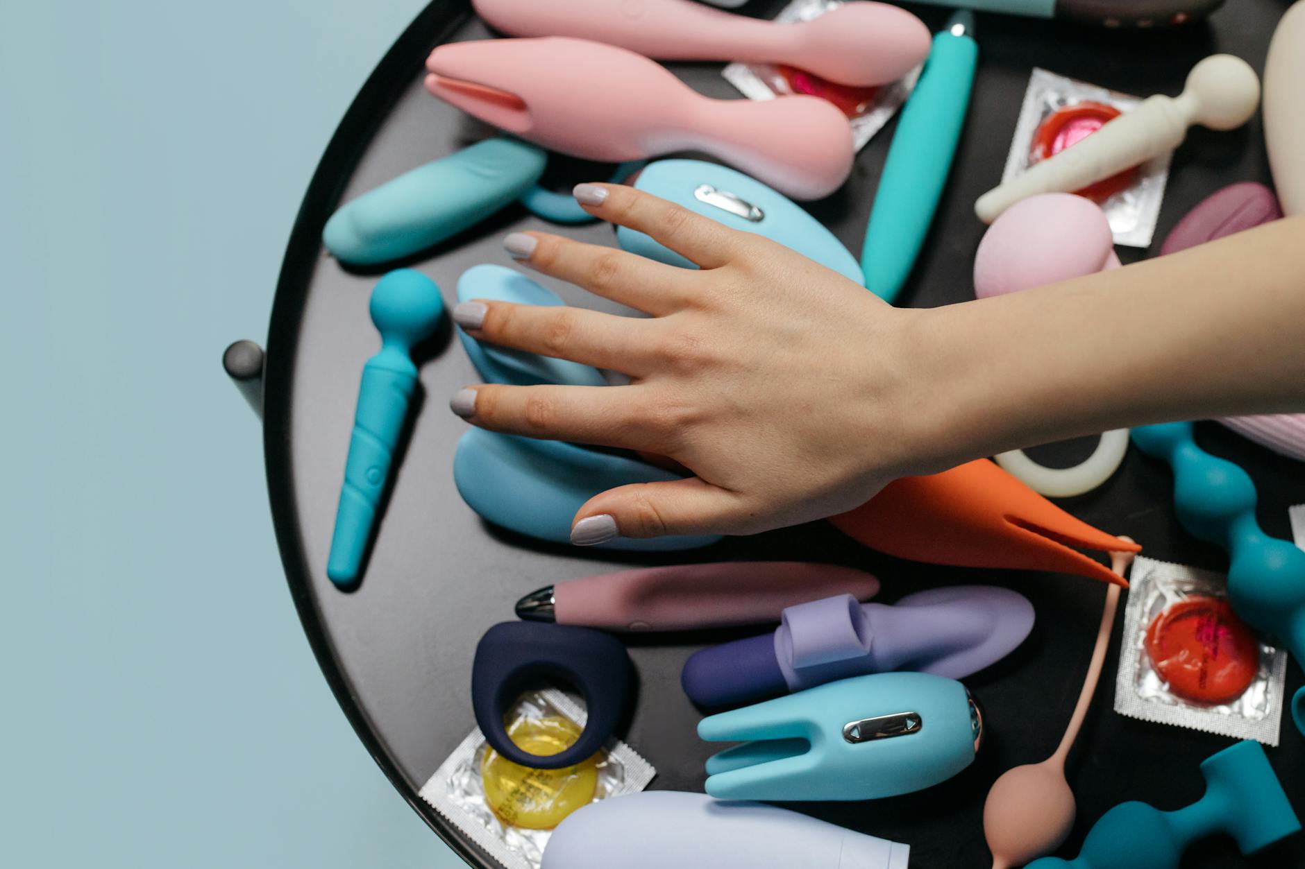 A hand reaching over a collection of colorful clitoral vibrators arranged on a table